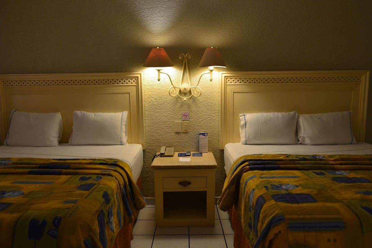 A hotel room with two beds, a nightstand with a phone and a lamp, and a textured wall.