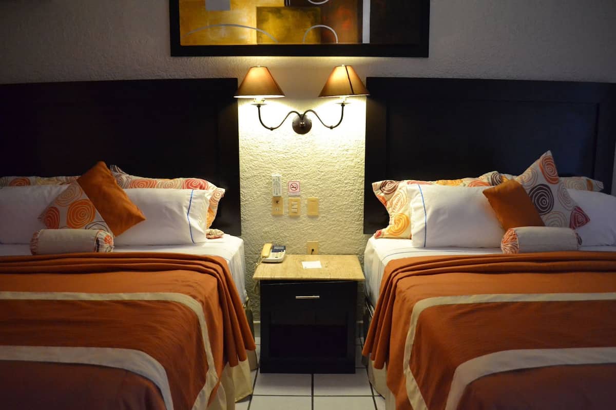 A hotel room with two beds, each with patterned pillows and orange bedding. There are two wall-mounted lamps above the beds, and a small bedside table with a phone and a notepad.