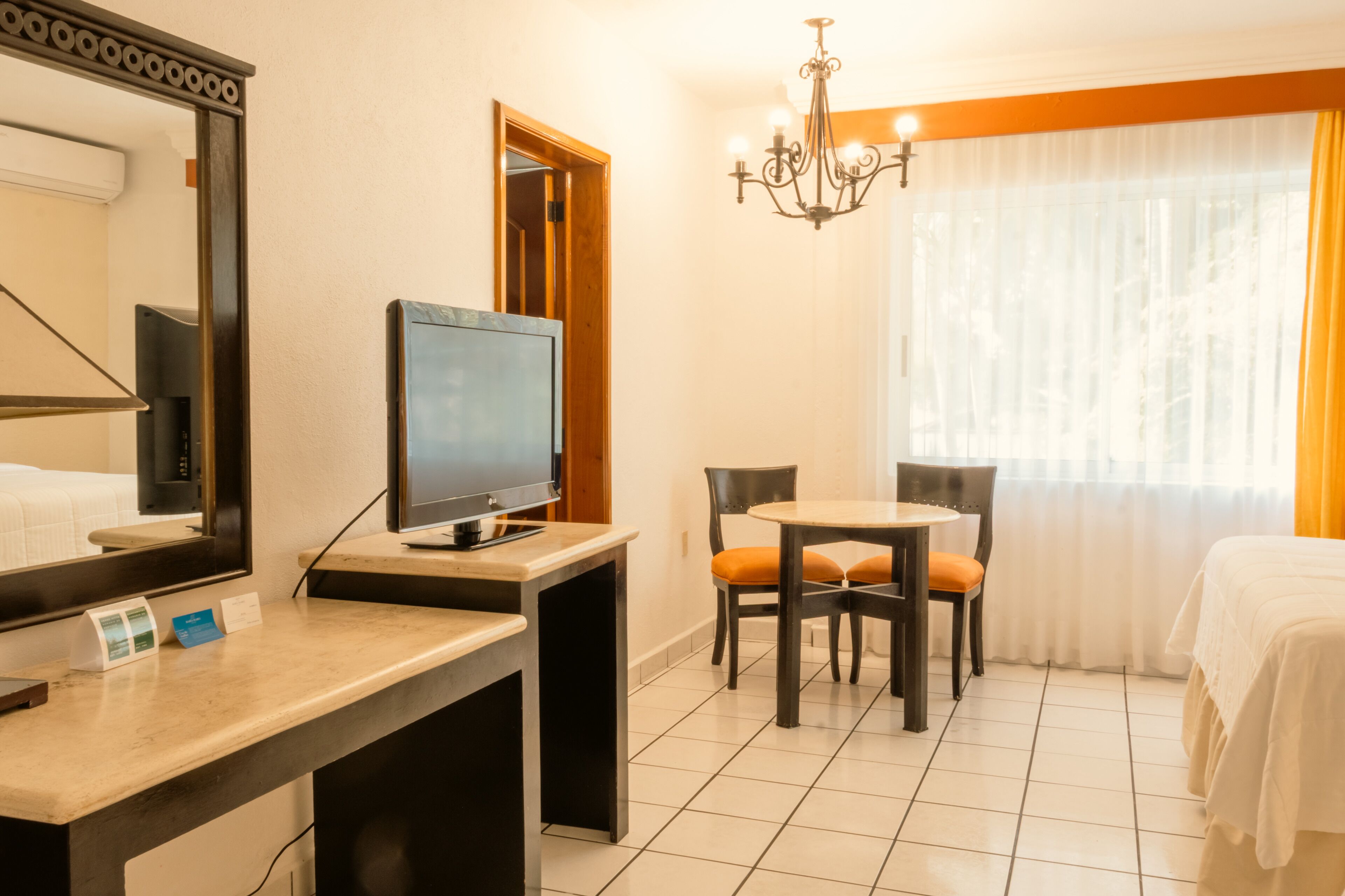 Una camera d'hotel con un letto, una scrivania con un computer, una sedia, un tavolo, un lampadario e una finestra con tende.