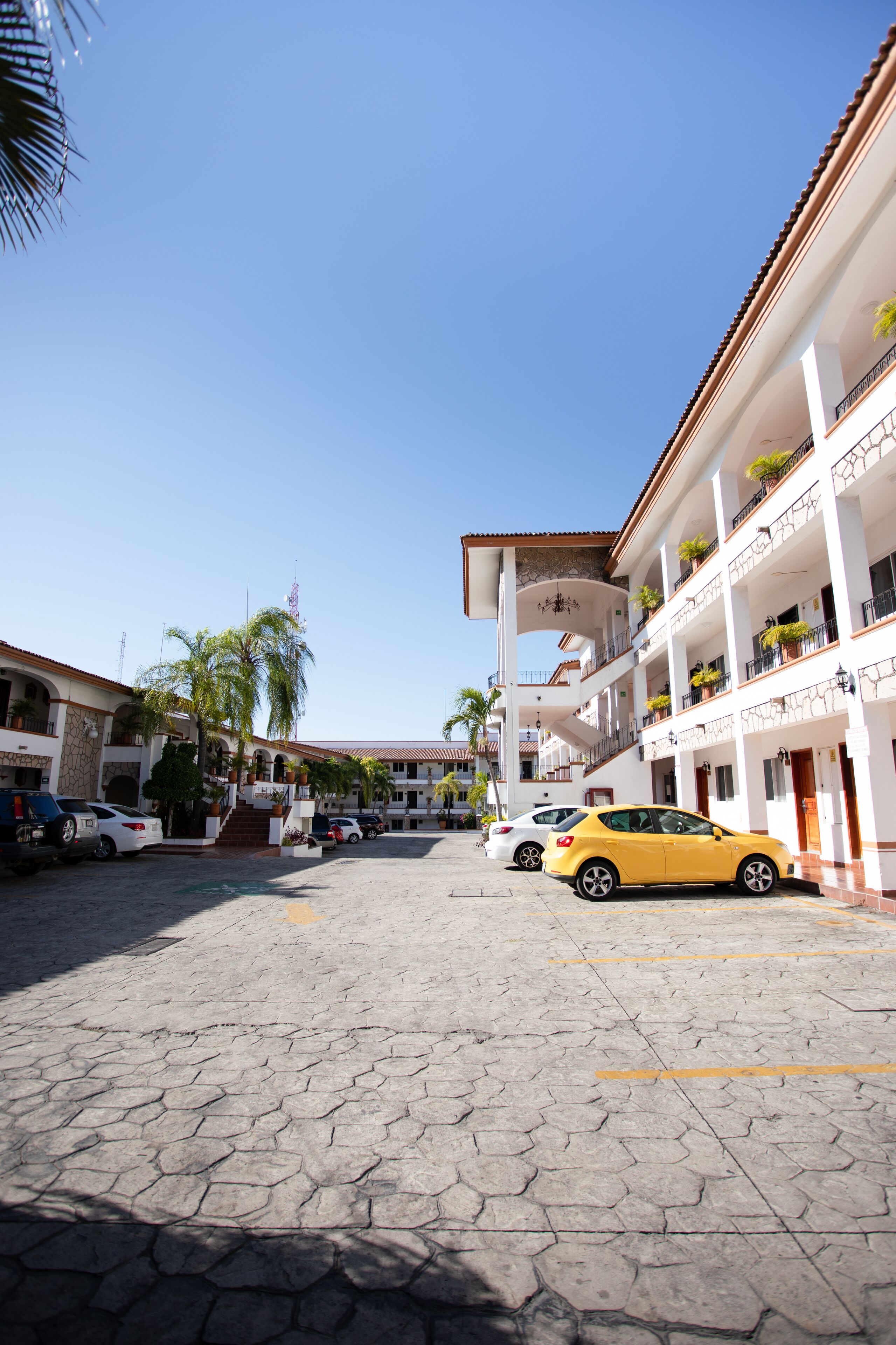 Um pátio com piso de azulejos, carros estacionados e um edifício com varandas e plantas em vasos.