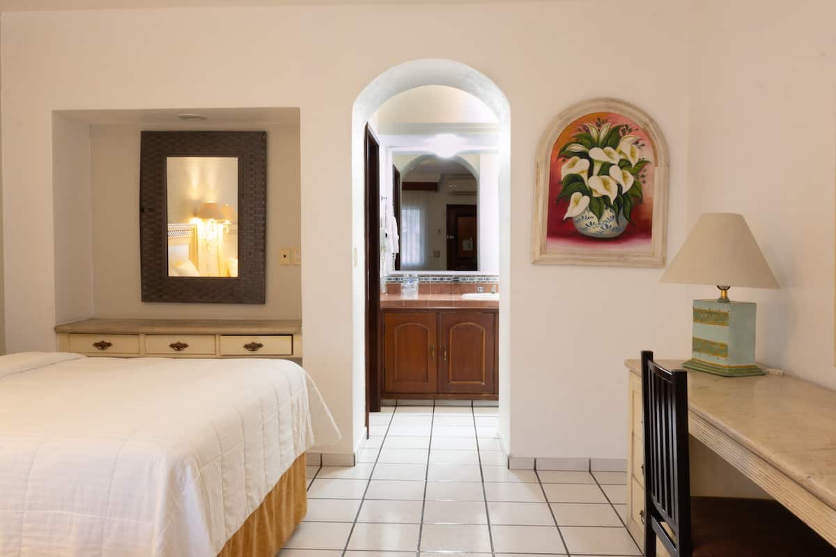 A hotel room with a bed, a desk, a lamp, a mirror, and a painting of flowers.