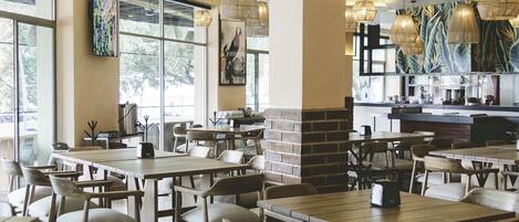 A spacious restaurant with high ceilings, modern pendant lighting, and large windows allowing natural light. Tables and chairs are arranged for dining.