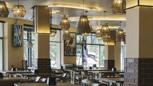 A spacious restaurant with multiple dining tables and chairs, large pendant lights, and a view of the outdoors through large windows.