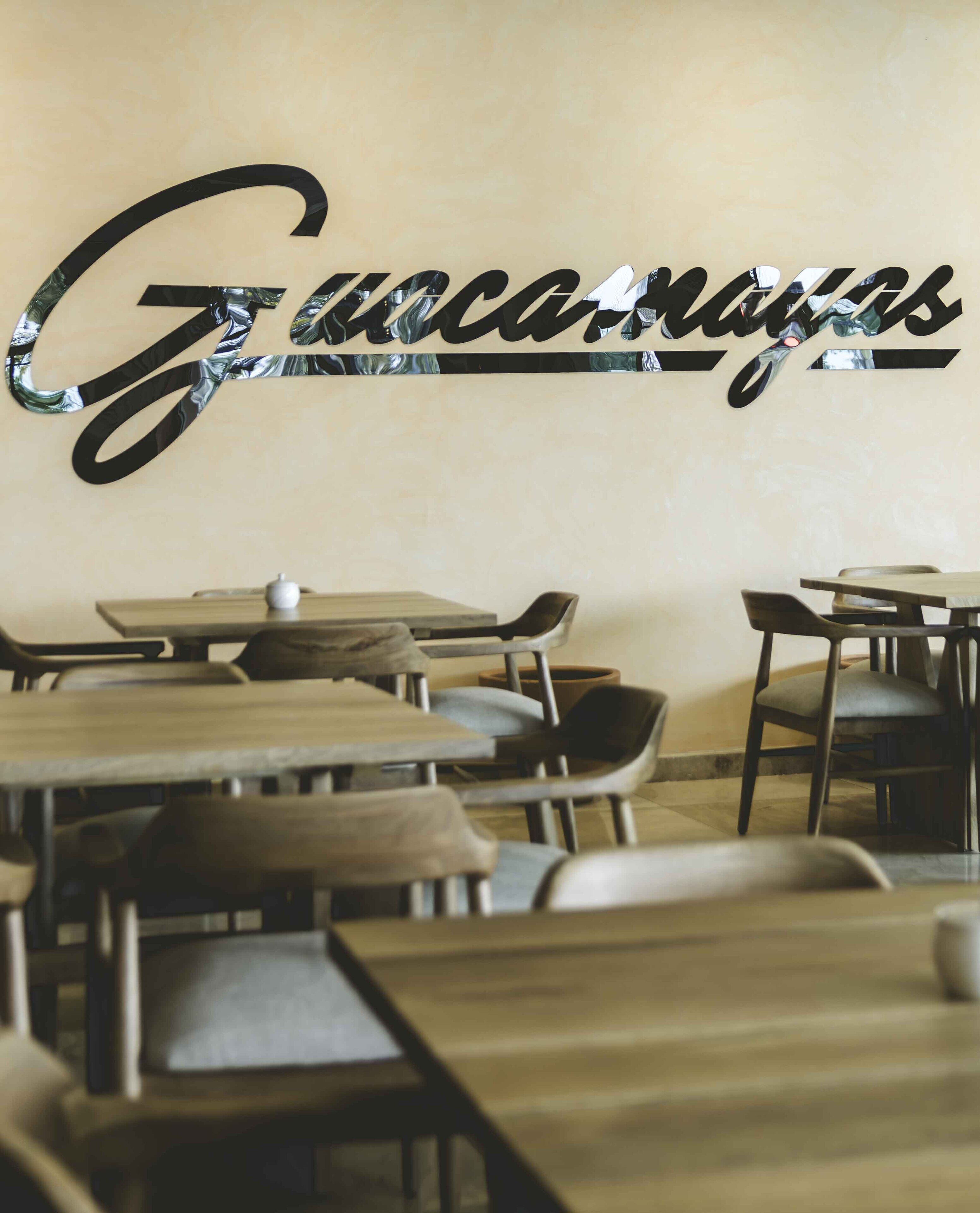 A restaurant interior with tables and chairs, a wall with the name 'Guacamex' in a stylized font, and a small white object on one table.