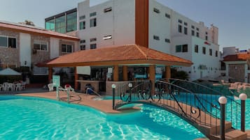 Piscina con sombrilla, zona de bar y vista a un edificio de varios pisos con balcones.