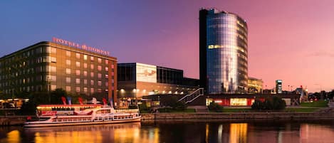 A hotel with a modern glass facade, a boat docked in front, and a river reflecting the buildings.