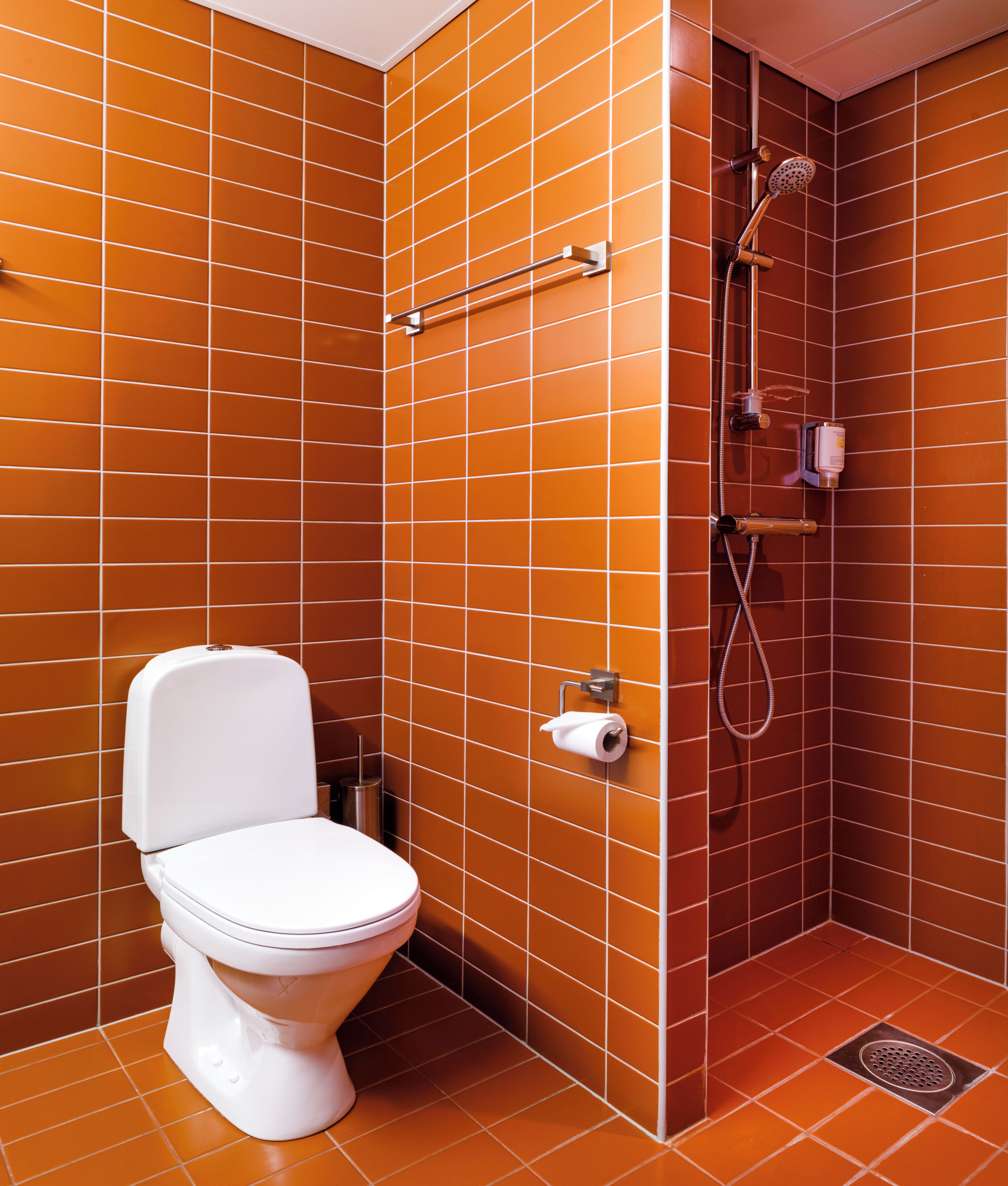 Une salle de bain avec des murs recouverts de carreaux orange, des toilettes blanches, une douche avec un pommeau de douche amovible et un porte-serviettes.
