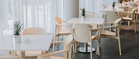 Un comedor moderno con mesas y sillas blancas, ventanas amplias con cortinas translúcidas y un piso de baldosas impecable.