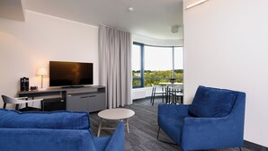 Una habitación de hotel moderna con una televisión de pantalla plana, una zona de comedor con mesa y sillas, un sofá y un amplio ventanal con vistas a zonas verdes.