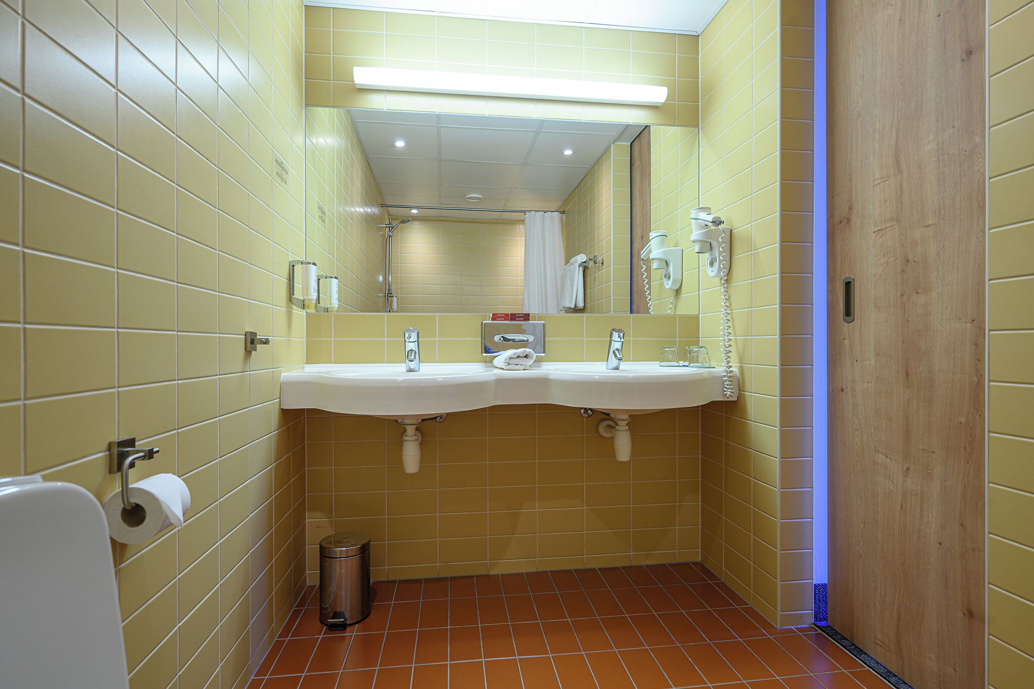 A bathroom with yellow tiled walls, a white sink with a mirror, a towel rack, and a trash can.