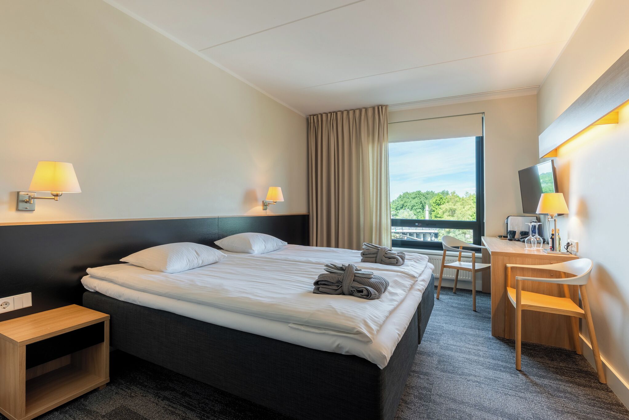 A hotel room with a bed, two bedside lamps, a desk with a chair, and a view of greenery outside.