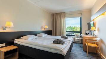 A hotel room with a bed, two bedside lamps, a desk with a chair, and a view of greenery outside.