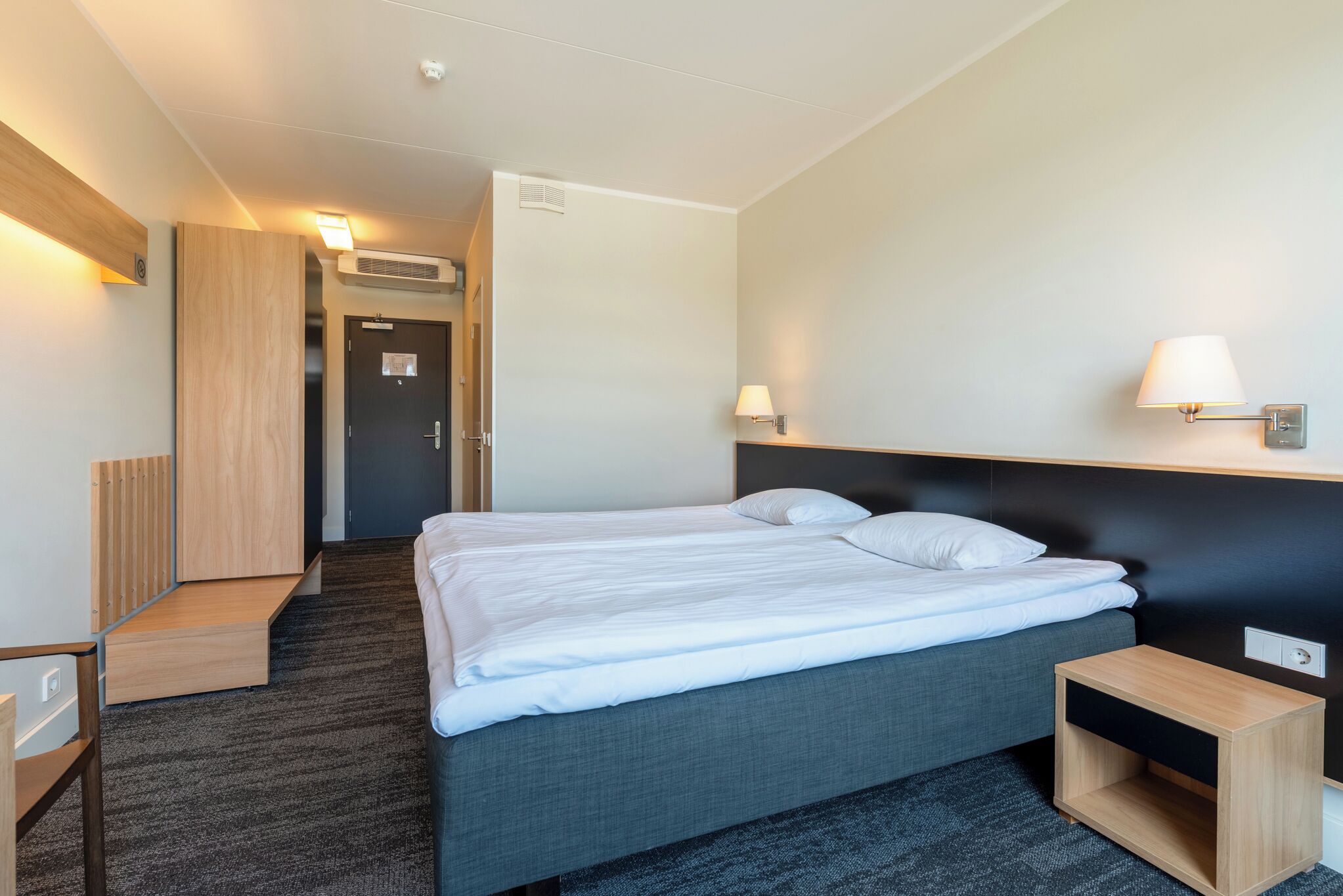 Una habitación de hotel con cama, mesitas de noche, armario y lámpara.