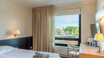 Quarto de hotel com cama, escrivaninha, cadeira, luminária, janela com vista para uma ponte e vegetação, e cortina.