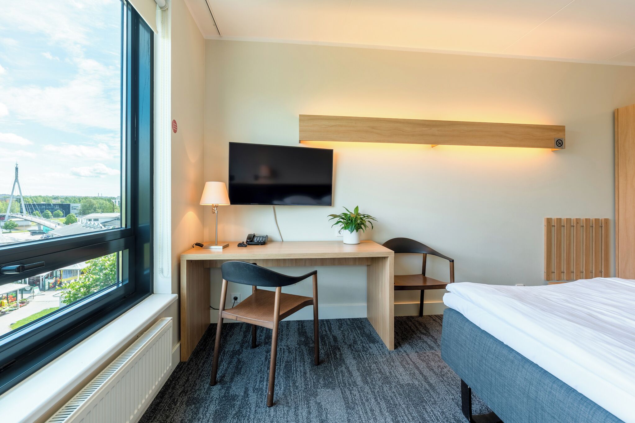 A hotel room with a desk, chair, bed, TV, and a window with a view of a bridge.