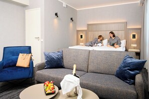 A modern hotel room with a sofa, a chair, a small table with a bottle and fruit, and a bed with two people. - Dorpat Hotel (Tartu)
