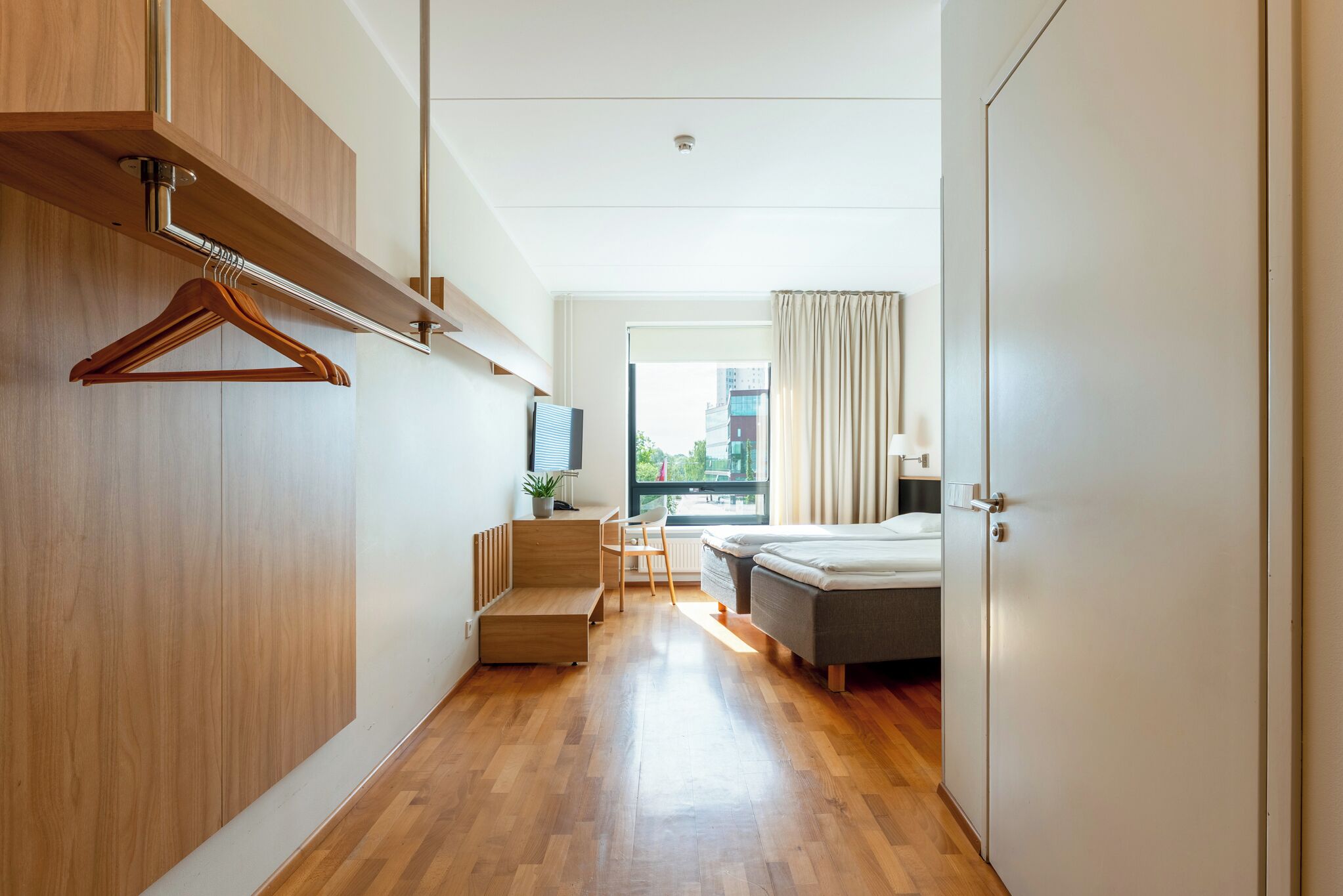 Habitación de hotel moderna con suelo de madera, cama, escritorio con silla, televisor y ventana con cortinas.