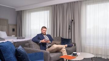 A man sitting on a grey sofa in a hotel room, using a laptop. There is a bed with a white mattress and grey pillows, a blue armchair, and a small round table with a cup and saucer.