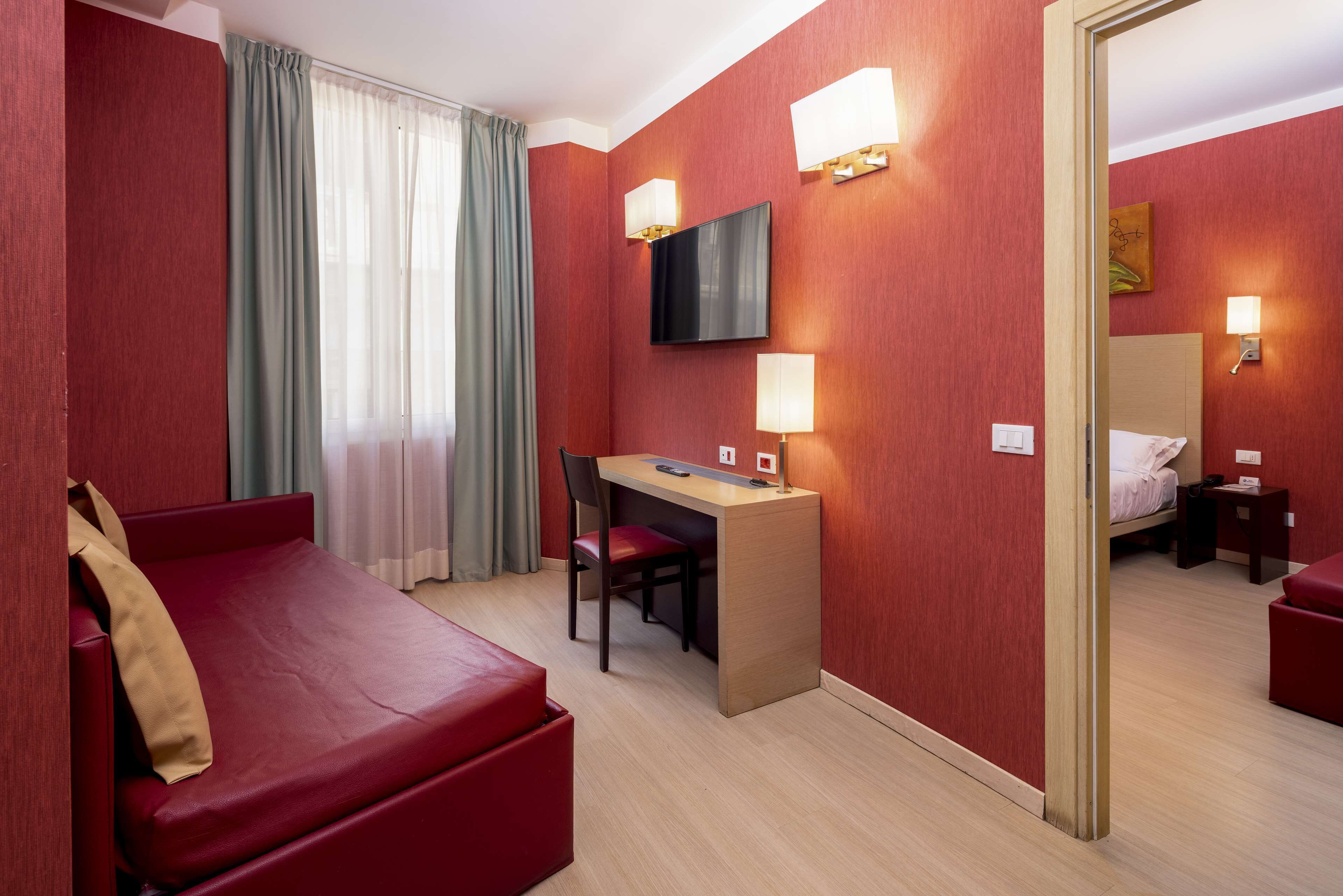 A hotel room with a red wall, a bed, a desk with a chair, a TV, and a mirror.