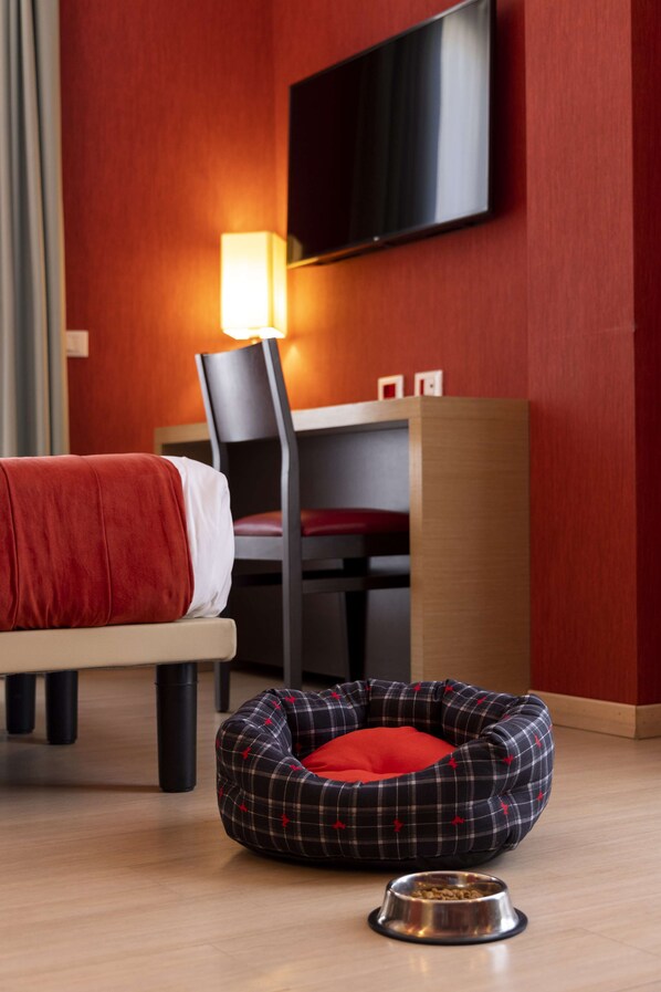 Une chambre d’hôtel comprenant un lit, un bureau, une chaise, un téléviseur fixé au mur et un panier pour animaux de compagnie avec un bol pour la nourriture.