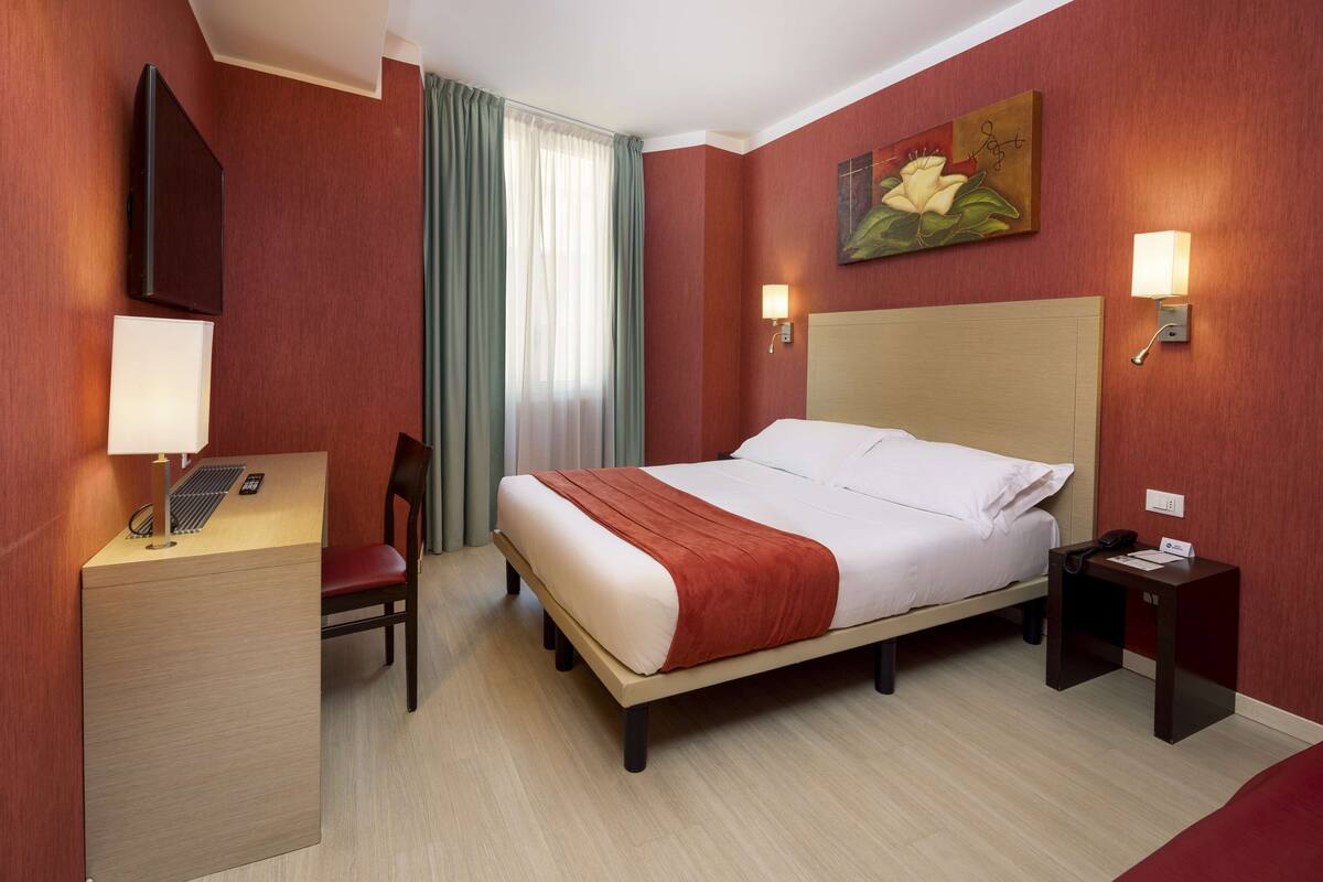A hotel room with a bed, desk, chair, and a painting on the wall.