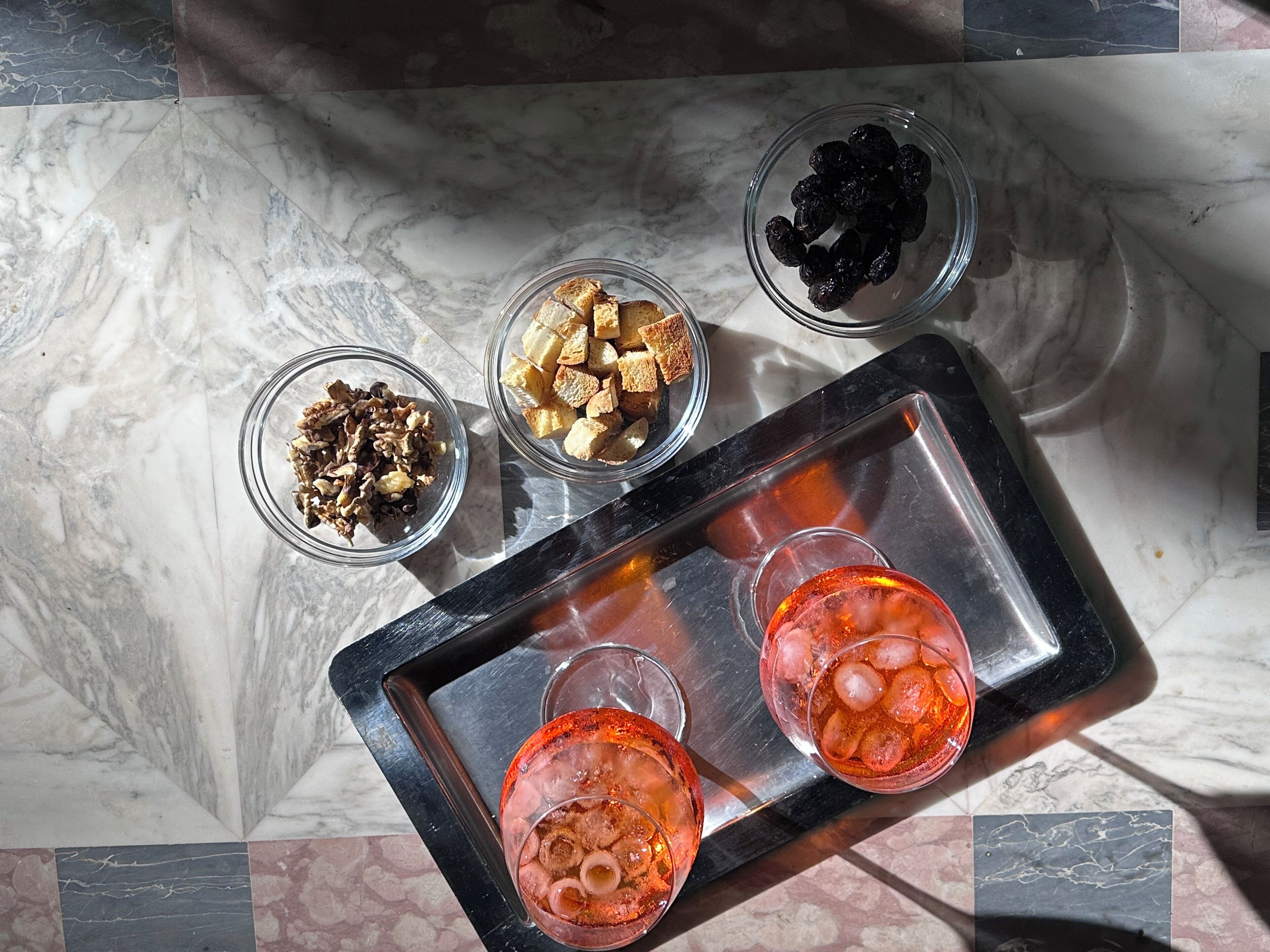 Una encimera de mármol con una bandeja que contiene dos vasos de una bebida roja con cubitos de hielo, un tazón de bayas oscuras, un tazón de granola y un tazón de cubos de pan.