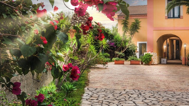 A courtyard with a paved walkway, a building with an arched entrance, and lush greenery including bougainvillea flowers. - Casale Romano Resort (Motta Camastra)