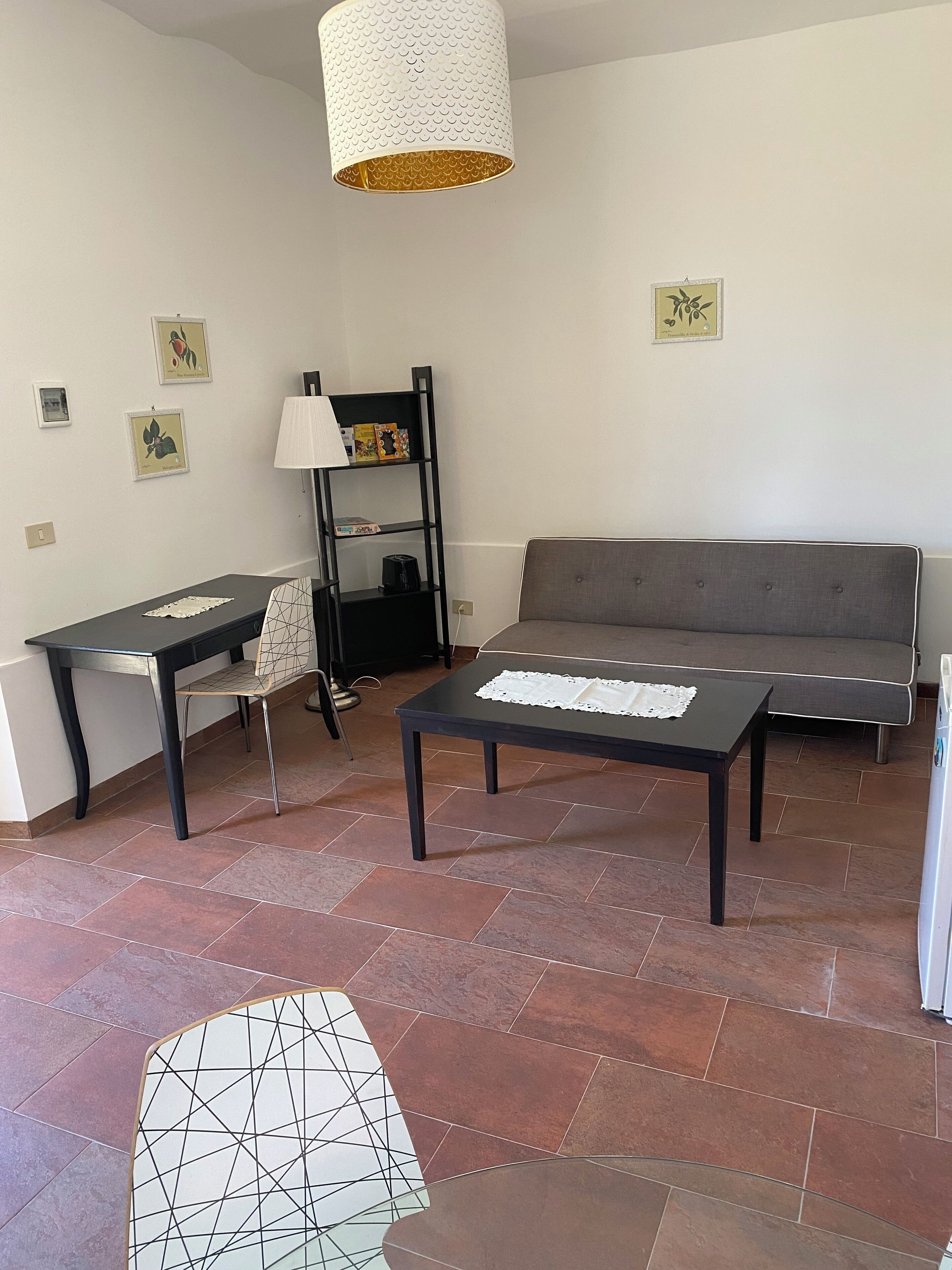 A living room with a sofa, a coffee table, a desk, and a chair. There is a bookshelf and a hanging light fixture.
