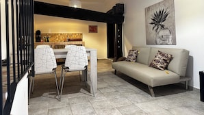 A modern living room with a sofa and a dining table with chairs. There is a kitchen in the background with a tiled backsplash and a hanging light fixture. - Casale Romano Resort (Motta Camastra)