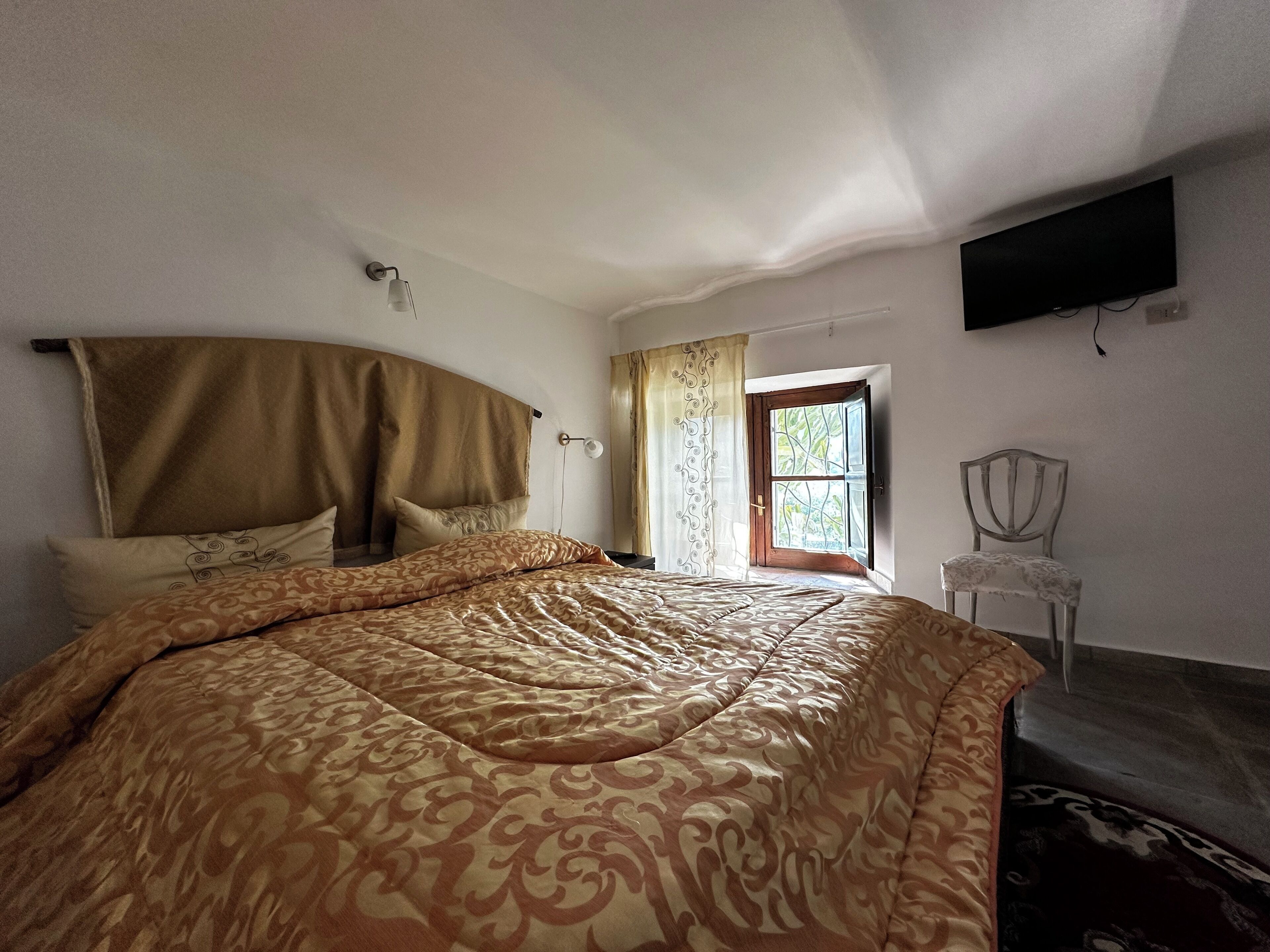 A bedroom with a large bed, a chair, a TV mounted on the wall, and a window with curtains.