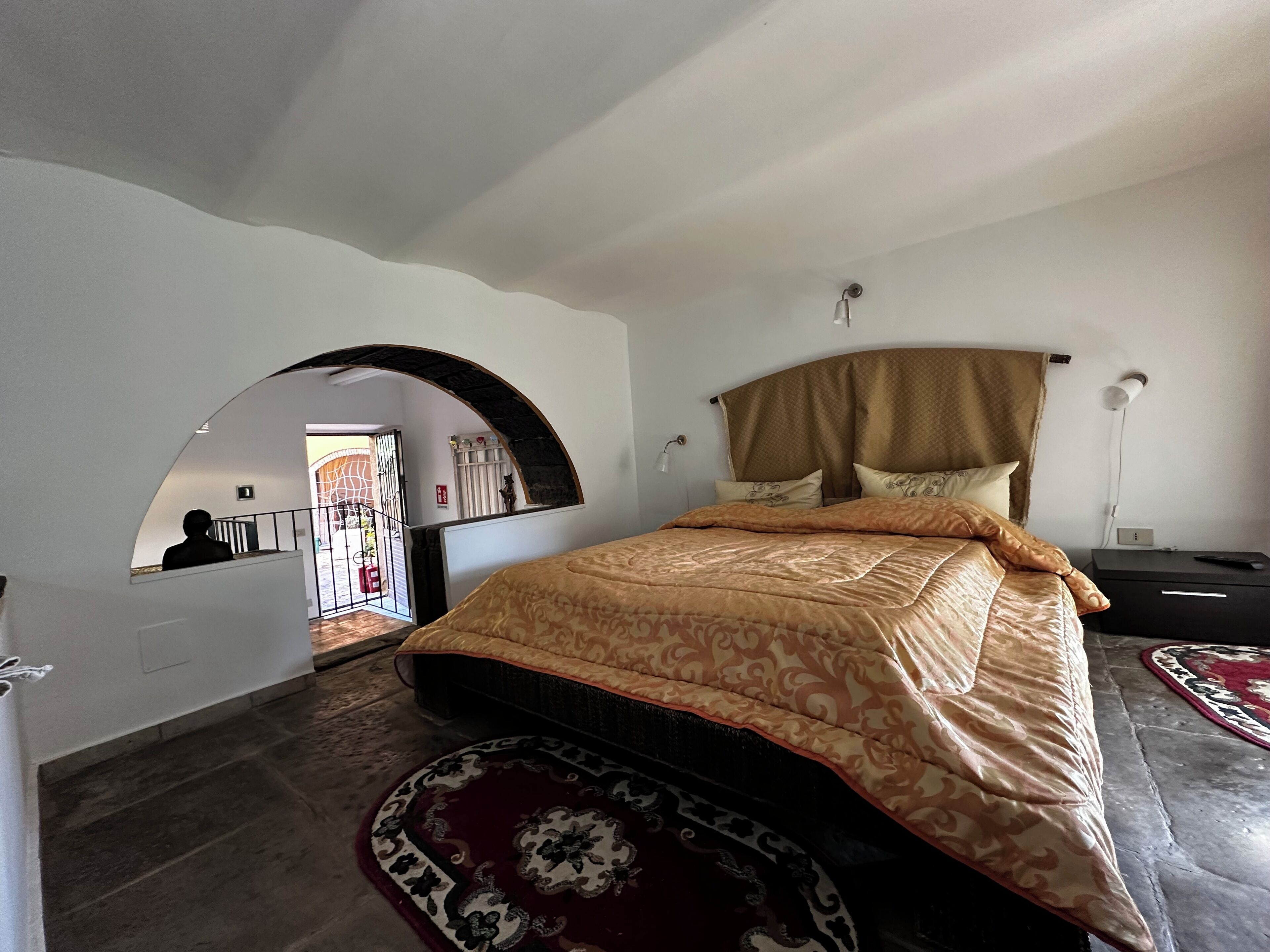 A bedroom with a large bed, a headboard, a nightstand, a lamp, and a view of a courtyard through an arched doorway.