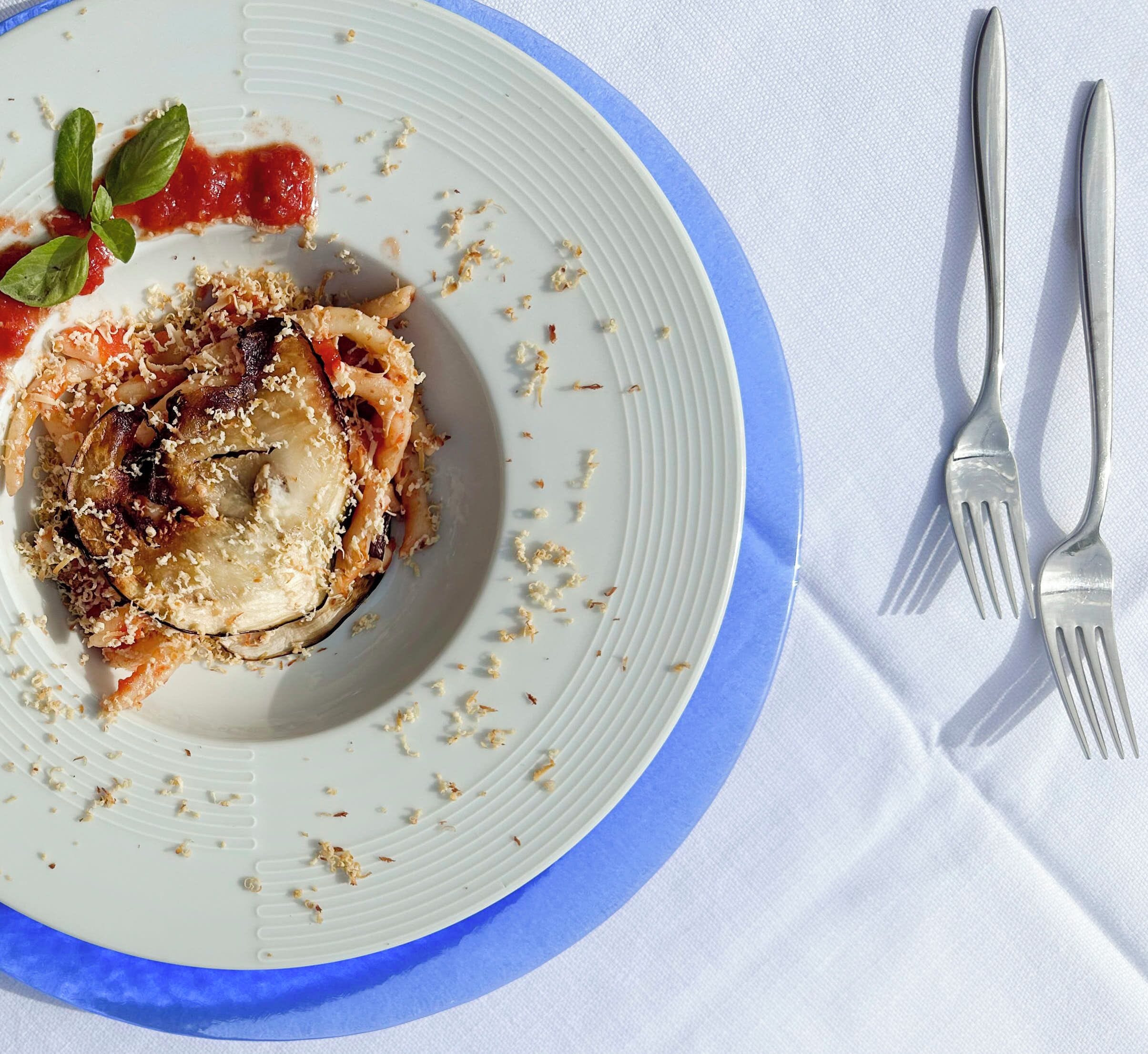 A plate of food with a blue rim, accompanied by two forks on a white tablecloth.