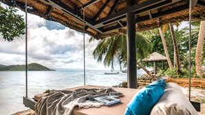 A beachside bed with a view of the sea, a boat in the distance, and a thatched-roof structure. - Matangi Island Resort - Adults Only (Matangi Island)