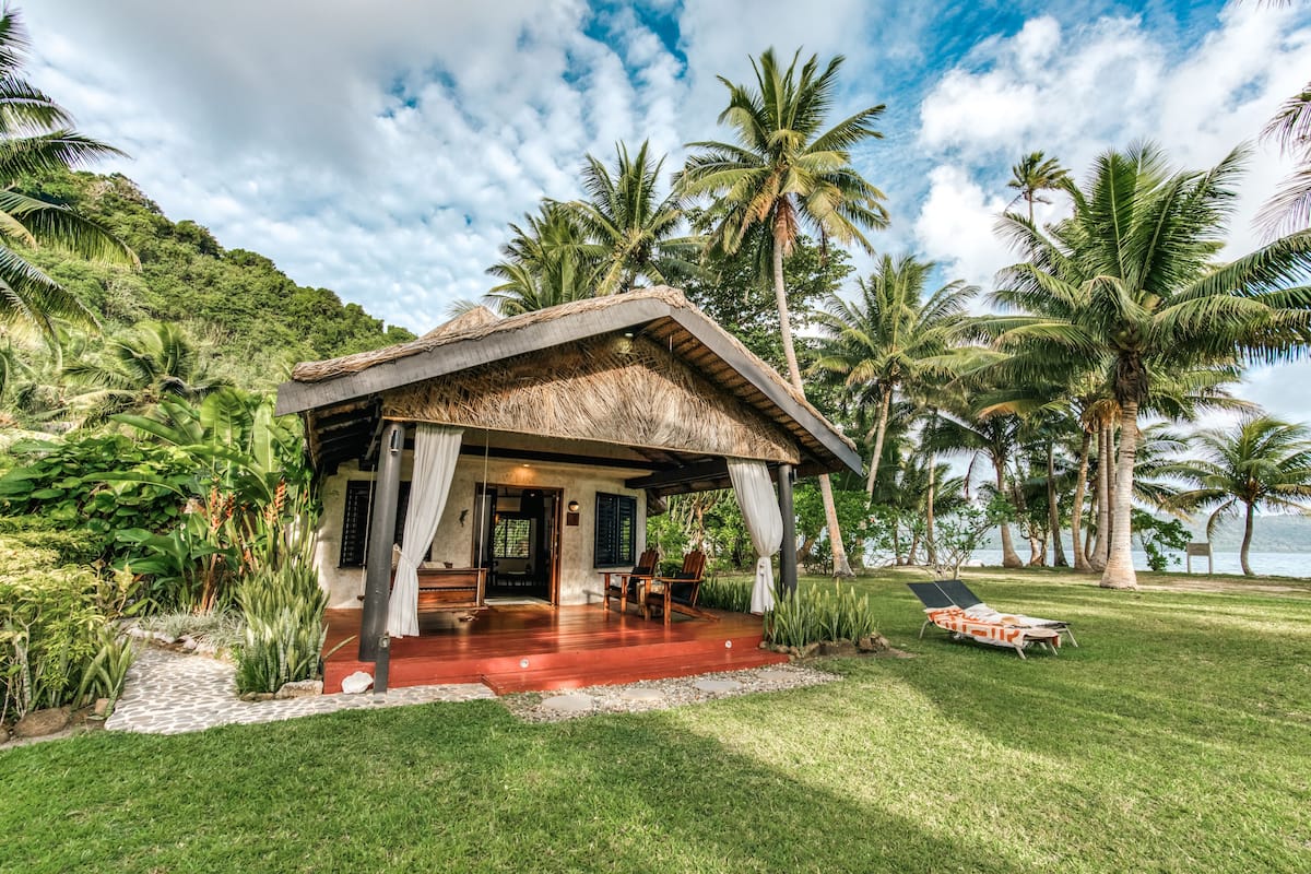 A small, thatched-roof structure with a covered porch, surrounded by lush greenery and palm trees, overlooking a body of water.