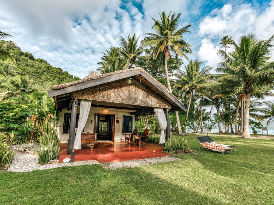 A small, thatched-roof structure with a covered porch, surrounded by lush greenery and palm trees, overlooking a body of water.