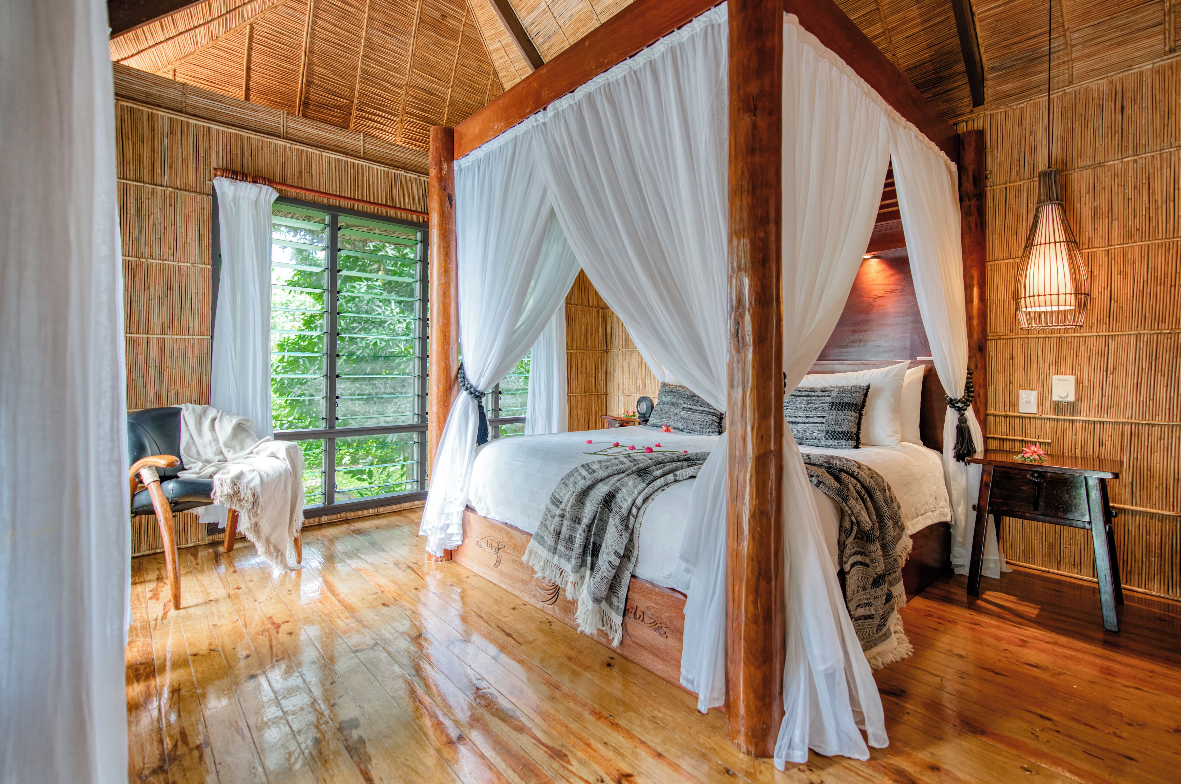 A four-poster canopy bed with a wooden floor, a bedside table with a lamp, and a view of greenery through large windows.