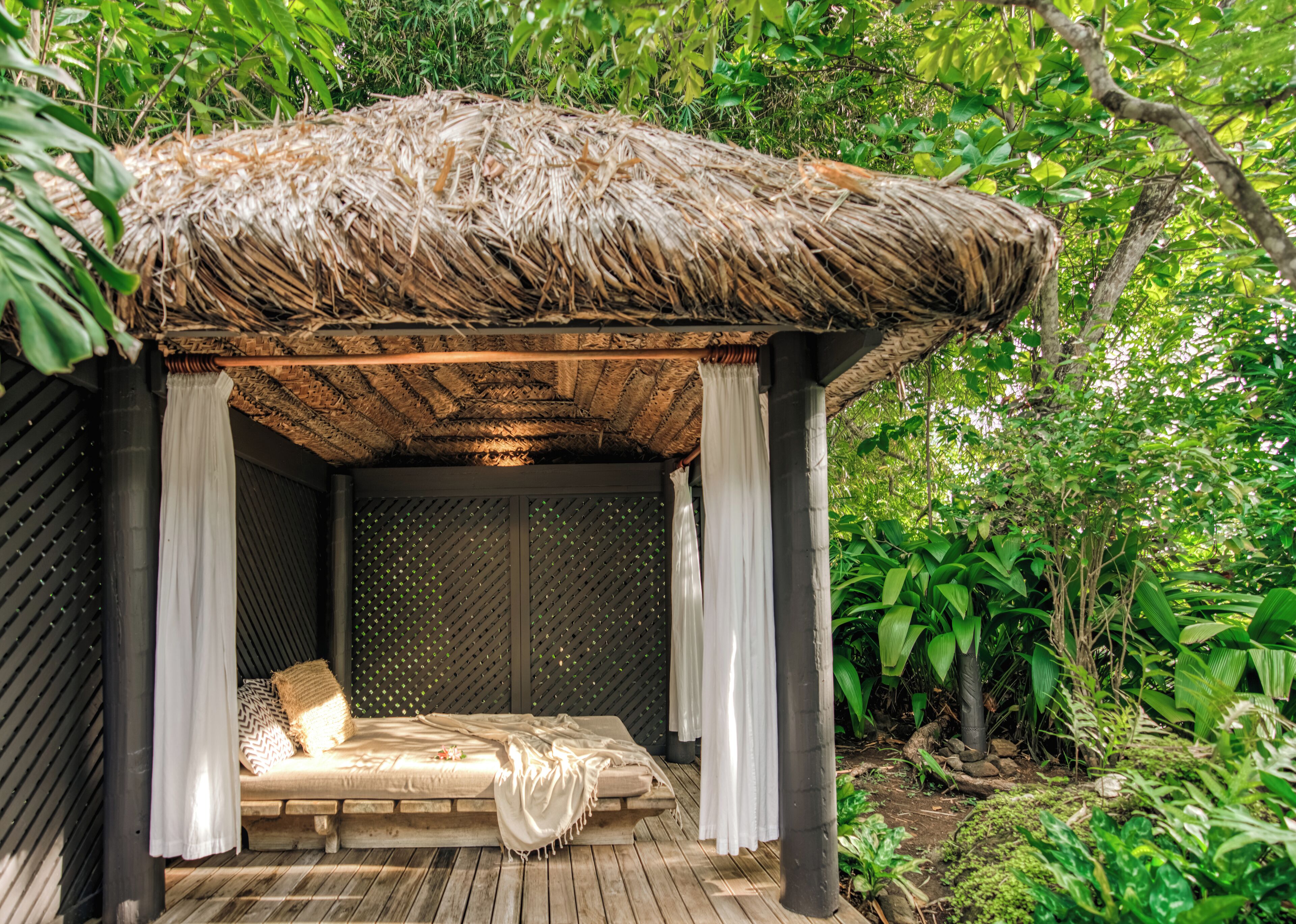 Un espacio para dormir al aire libre con techo de paja, cama, cortinas y una terraza de madera.