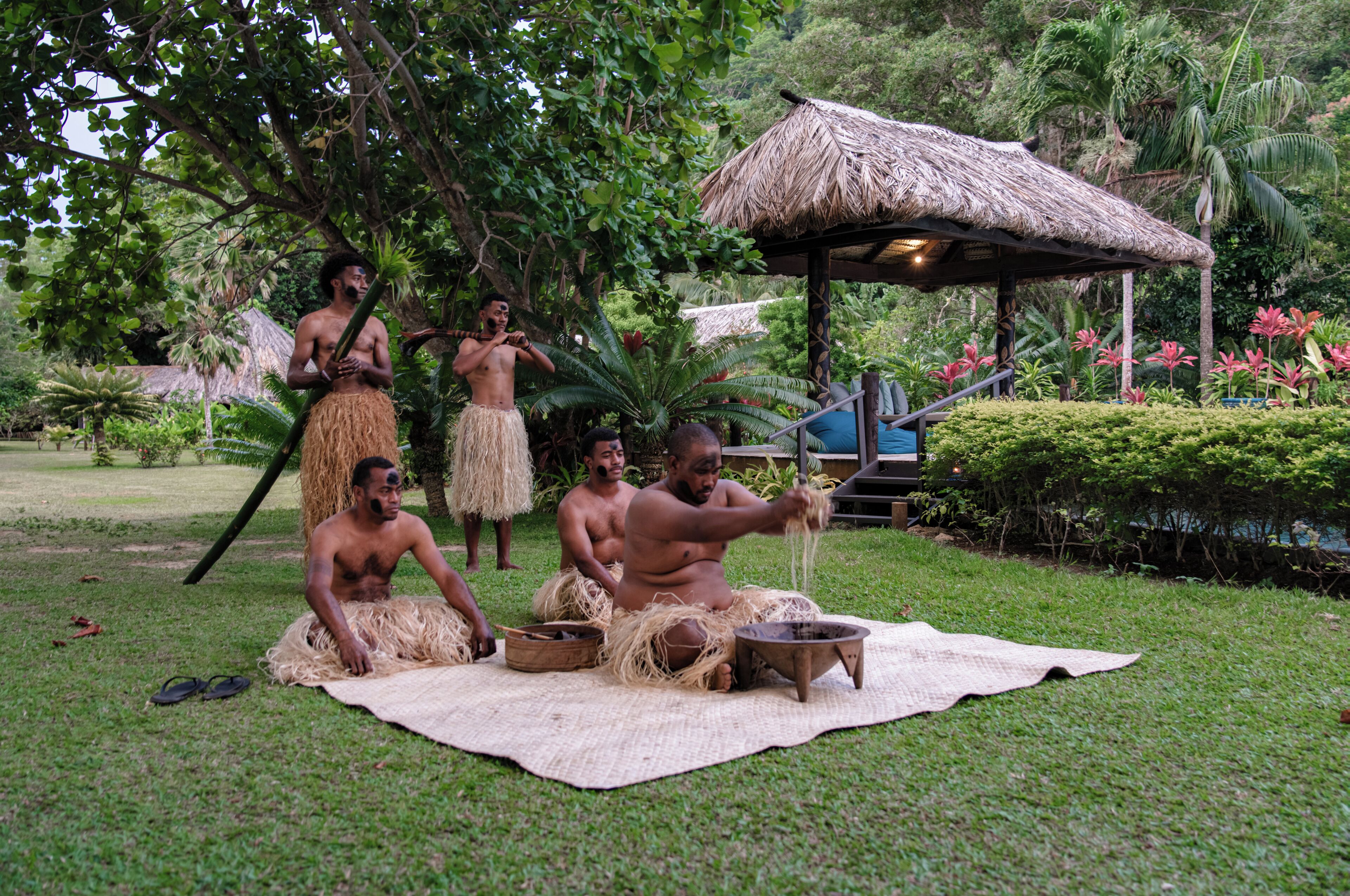 Um grupo de pessoas ao ar livre, envolvidas em alguma atividade, com uma estrutura de telhado de palha ao fundo.