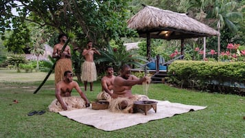 Um grupo de pessoas ao ar livre, envolvidas em alguma atividade, com uma estrutura de telhado de palha ao fundo.