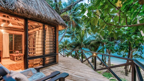 A wooden deck with a thatched-roof structure, a dining area with a table and chairs, and a view of the beach and palm trees. - Matangi Island Resort - Adults Only (Matangi Island)