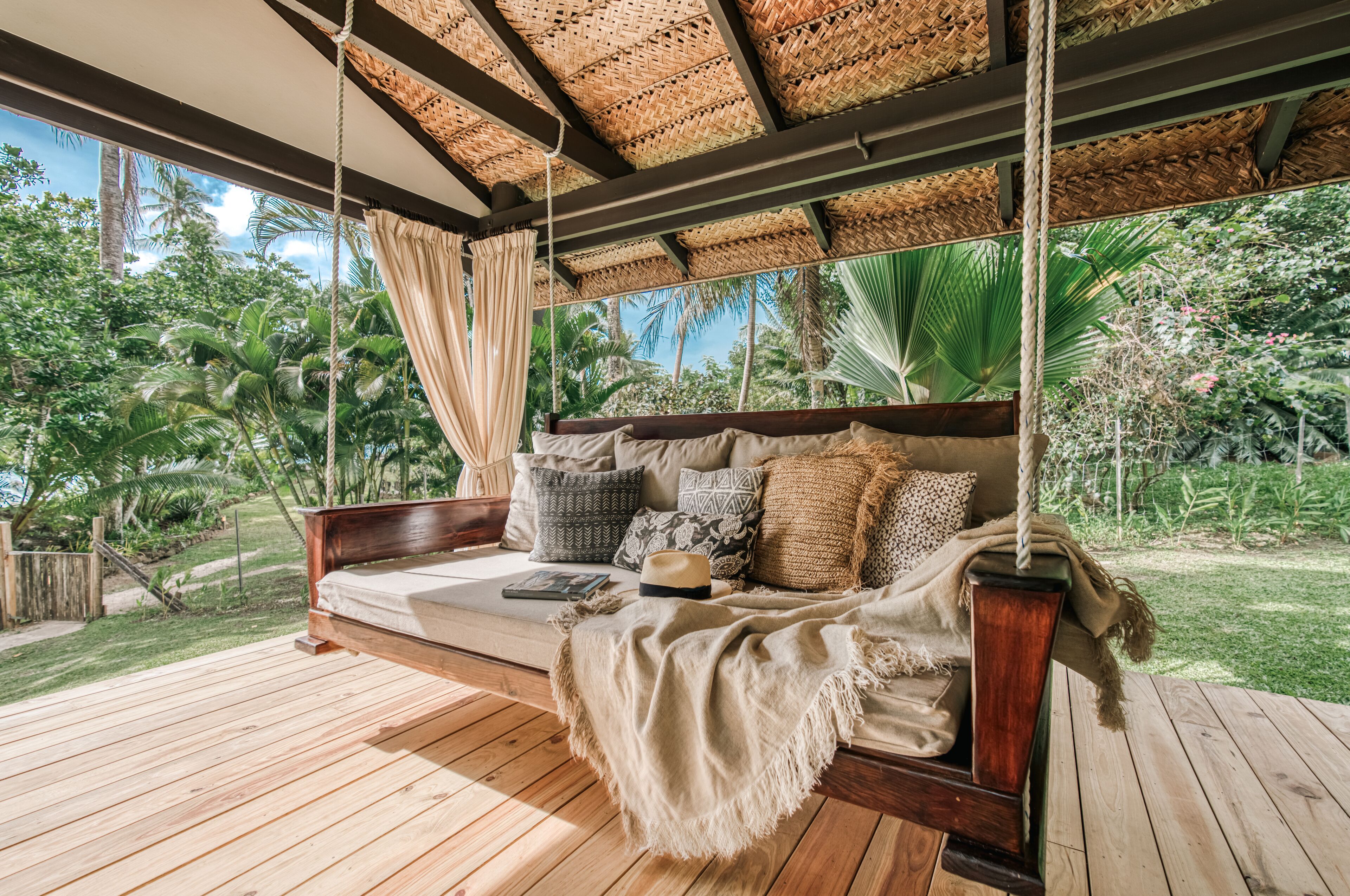 A wooden deck with a hanging swing bed, surrounded by lush greenery and a thatched roof.