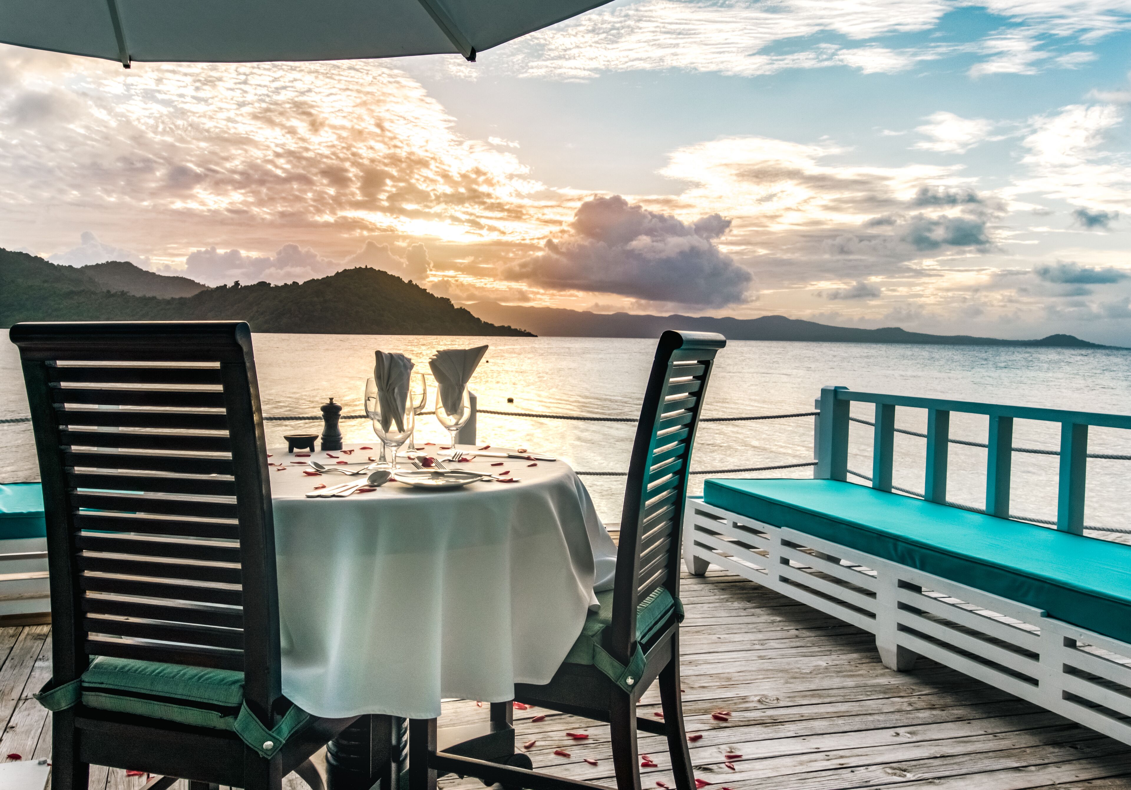A table set for two with chairs, overlooking a body of water and hills during sunset.