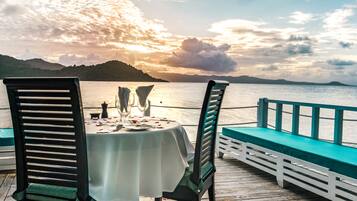 A table set for two with chairs, overlooking a body of water and hills during sunset.