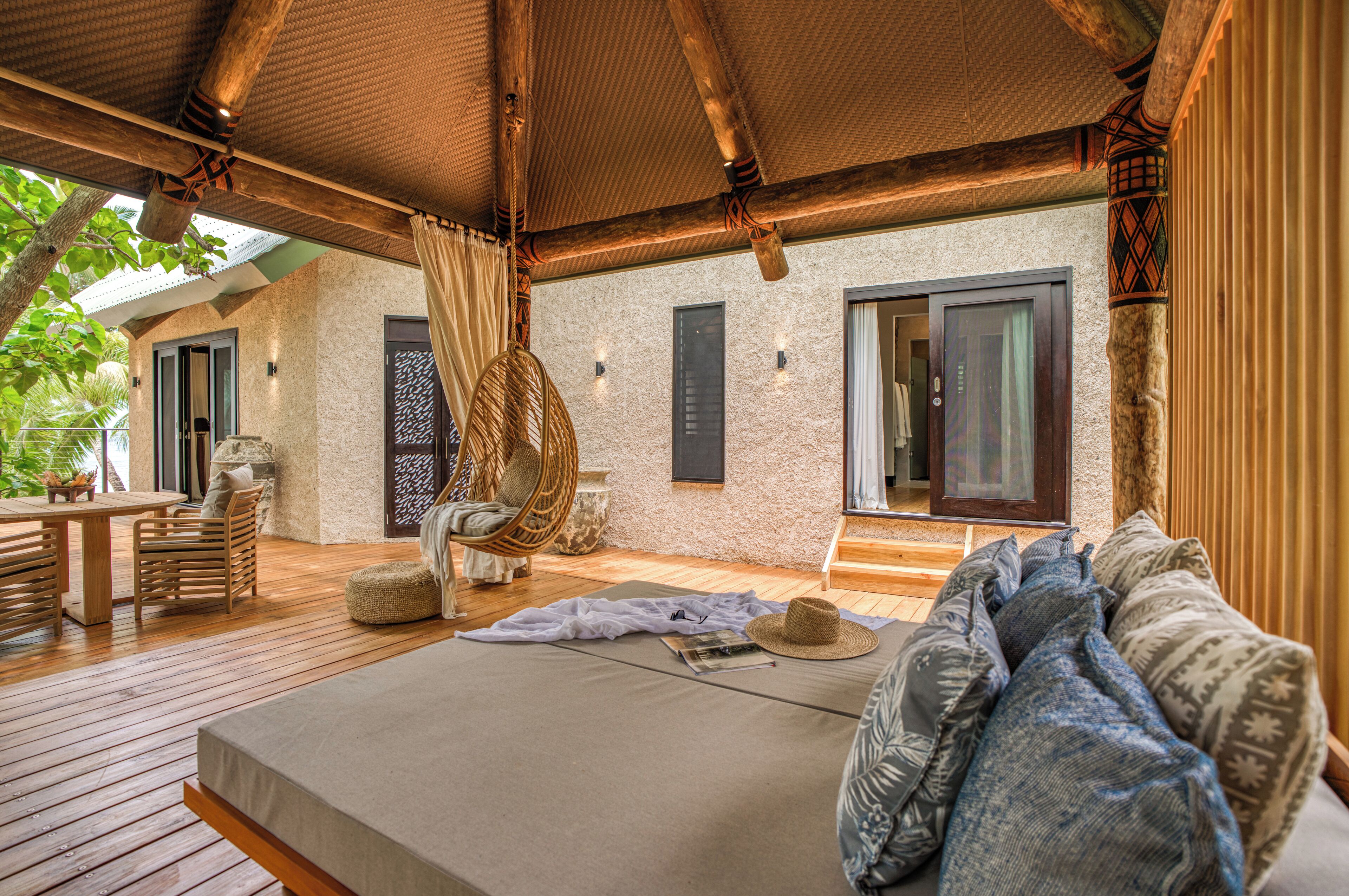 A spacious bedroom with a large bed, a wooden deck, wicker furniture, and a stone wall.