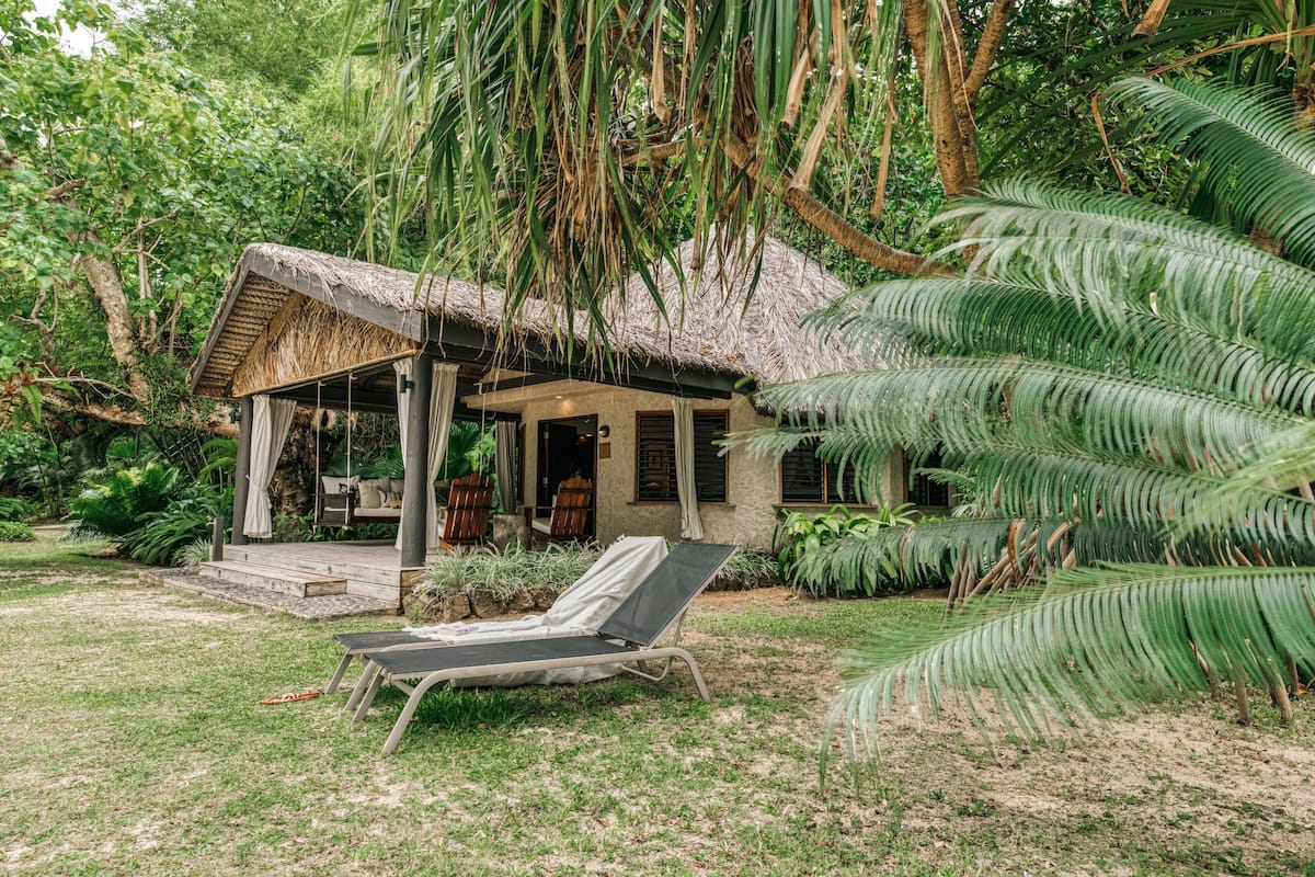 A thatched-roof hut with a covered porch, a lounge chair, and lush tropical vegetation.