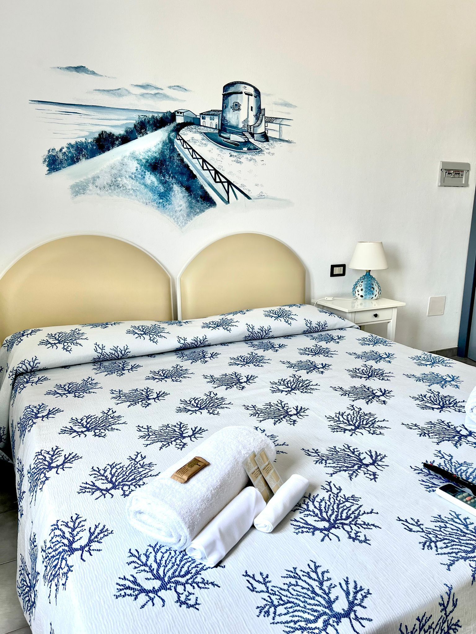 Habitación de hotel con cama que presenta un estampado azul y blanco de coral, una lámpara blanca sobre una mesita de noche y un mural en la pared con una escena costera.