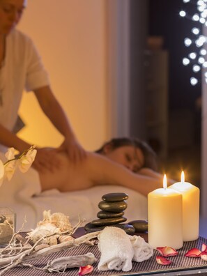 A massage therapy room with a person receiving a back massage, candles, and spa amenities like stones and towels. - Hotel Cala di Seta (Calasetta)