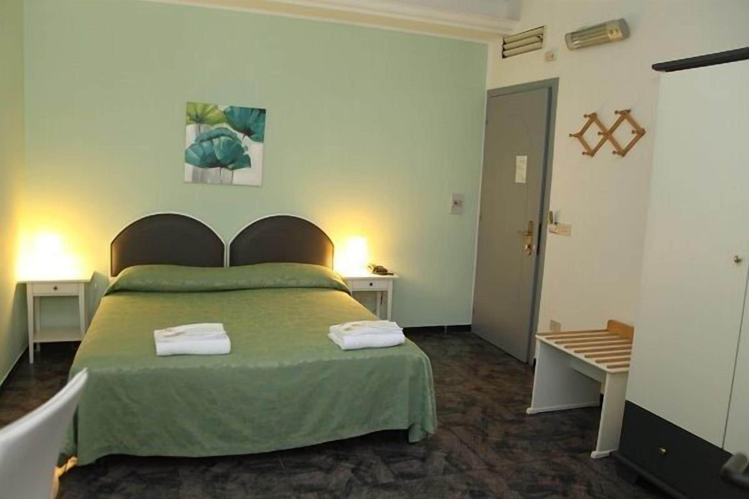 A hotel room with a bed, two bedside lamps, a green bedspread, a painting on the wall, a door, and a small bench.