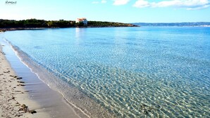 A beach with clear water, a sandy shore, and a building on a hill in the background. - Hotel Cala di Seta (Calasetta)