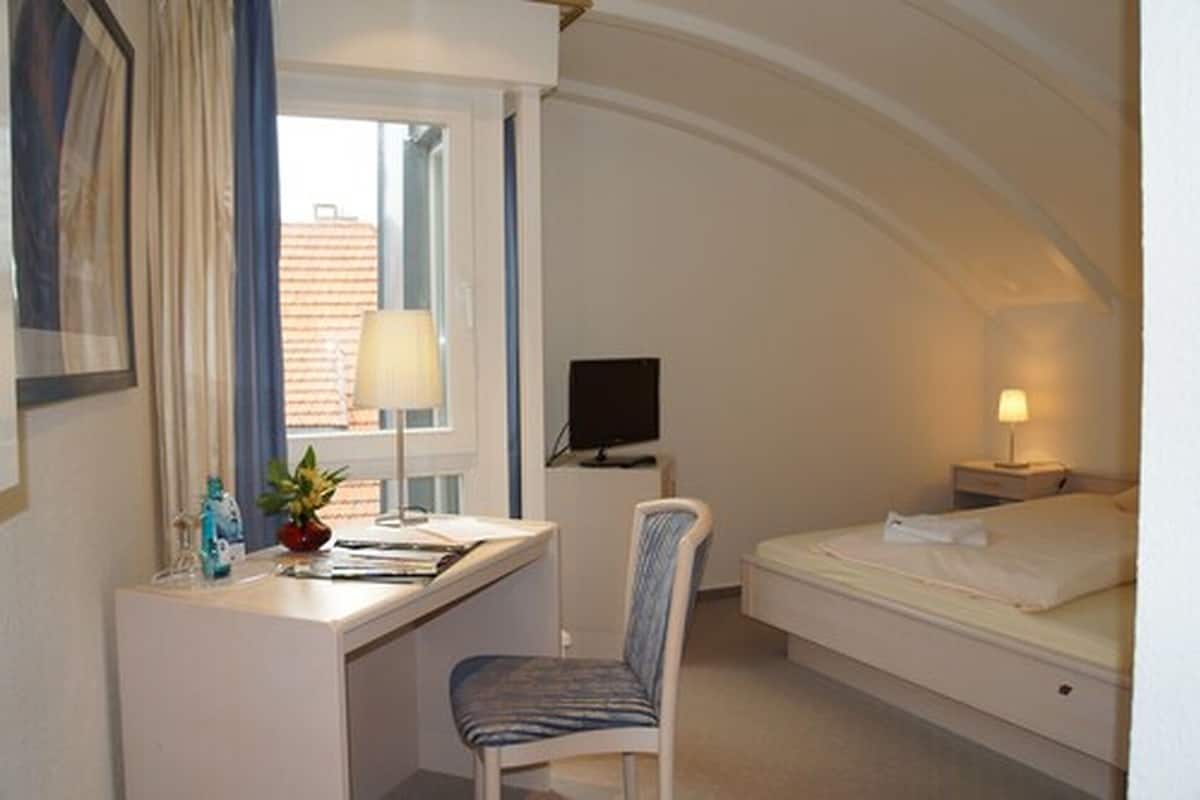 A compact hotel room with a desk, chair, bed, TV, and a window with curtains.